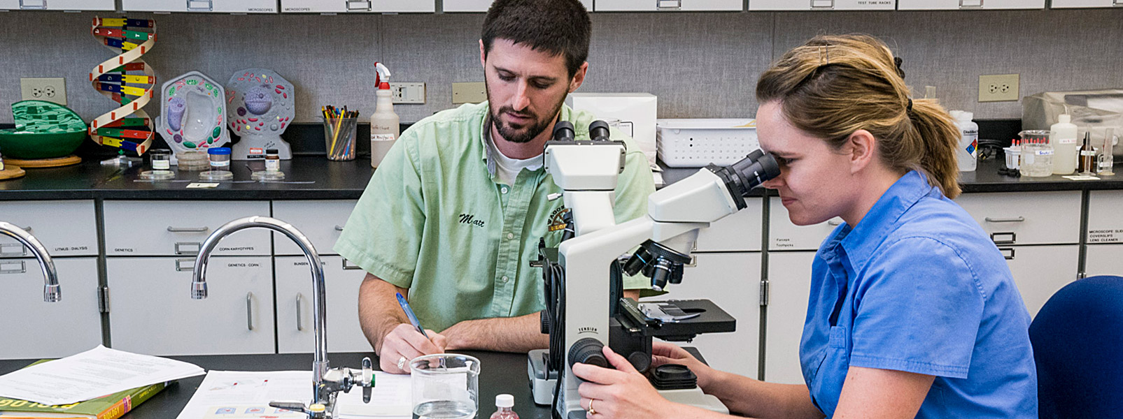 girl looking through microscope and guy taking notes